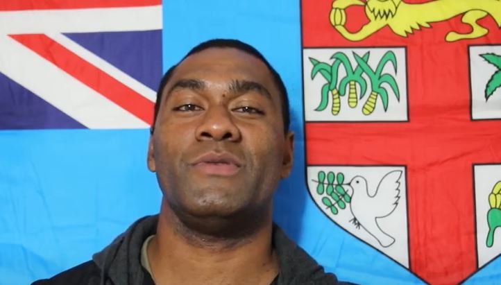 A man stands in front of a light blue Fijian flag.