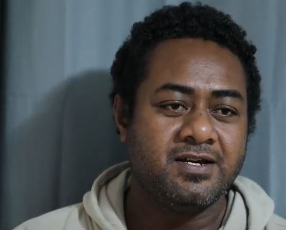 A Fijian man wearing a beige hoodie smiles as he talks to the camera.