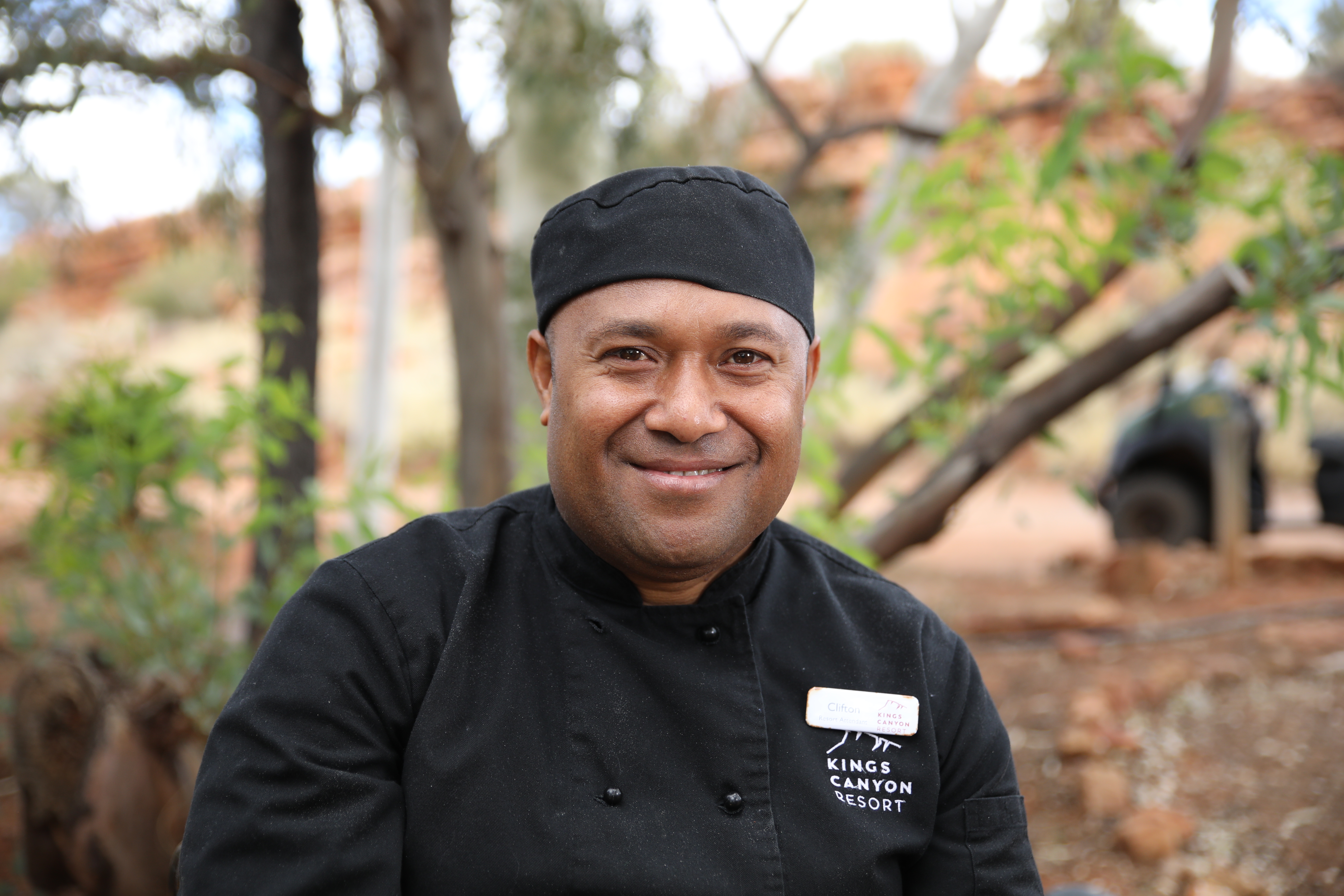 A chef in uniform smiling at the camera with some trees in the background