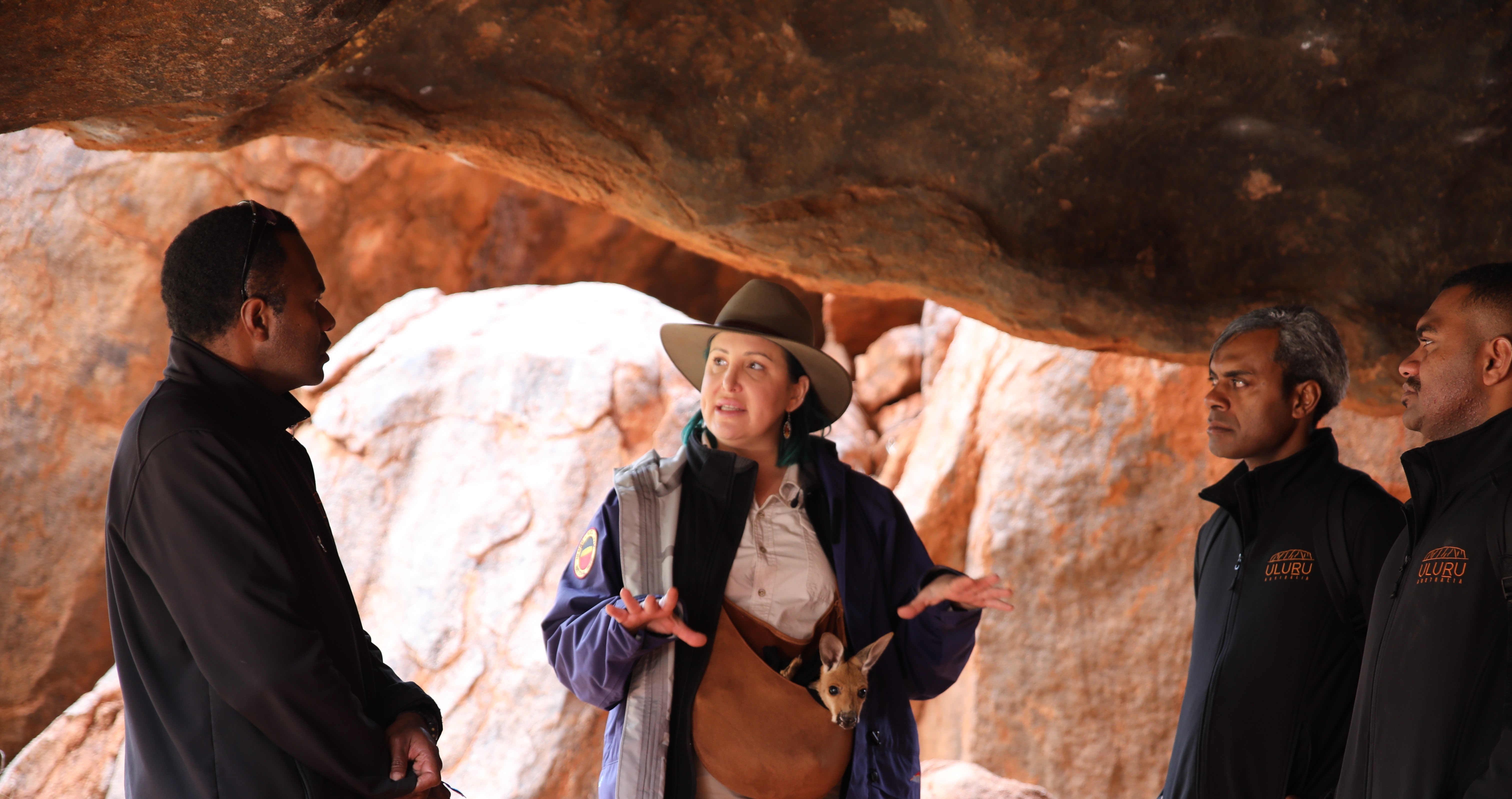 Three men stand talking with a women under a rock at Uluru 