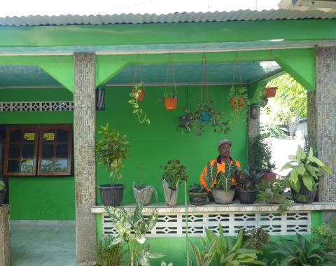 A Timorese man in an orange high-visibility shirt stands in front of a green house.