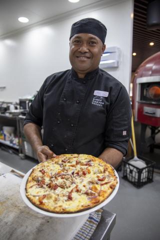 A chef standing in a kitchen holding a pizza and smiling at the camera 