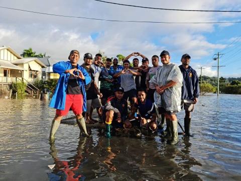 A group of men in long gumboots stand in ankle deep floodwaters, some wearing raincoats and some crouching. Some of the men make heart symbols with their hands as they all face the camera and smile.