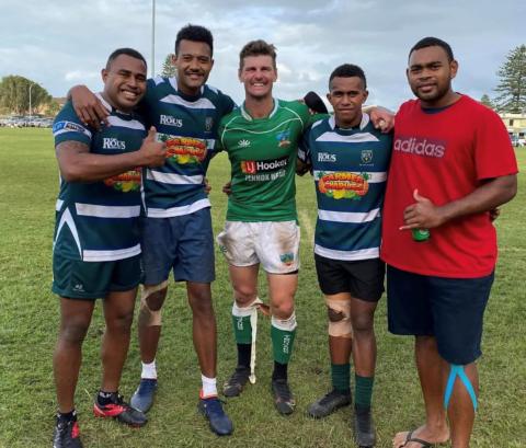 Five men wearing rugby jerseys and sports clothing stand smiling at the camera with their arms around each other.