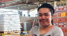 A woman wearing black gloves and a grey tshirt with her brown hair tied back smiles at the camera. She is standing in a packing shed with boxes behind her, filling out paperwork as she leans against a box.