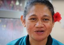 A Samoan woman wearing a blue shirt and a red flower in her hair smiles.