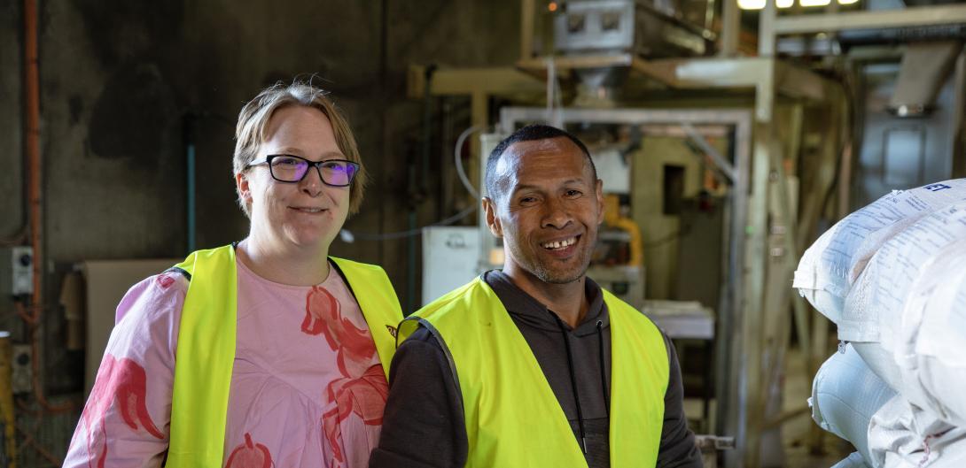 A man and a woman stand smiling at the camera wearing high vis vests in a warehouse 